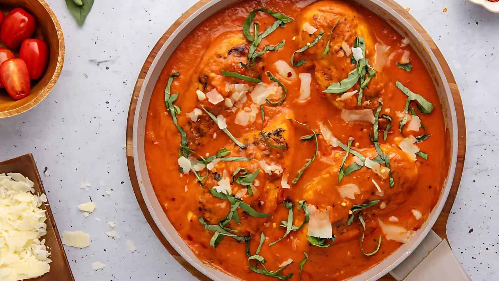 A skillet filled with chicken breasts in tomato-based sauce, garnished with shredded cheese and fresh basil.