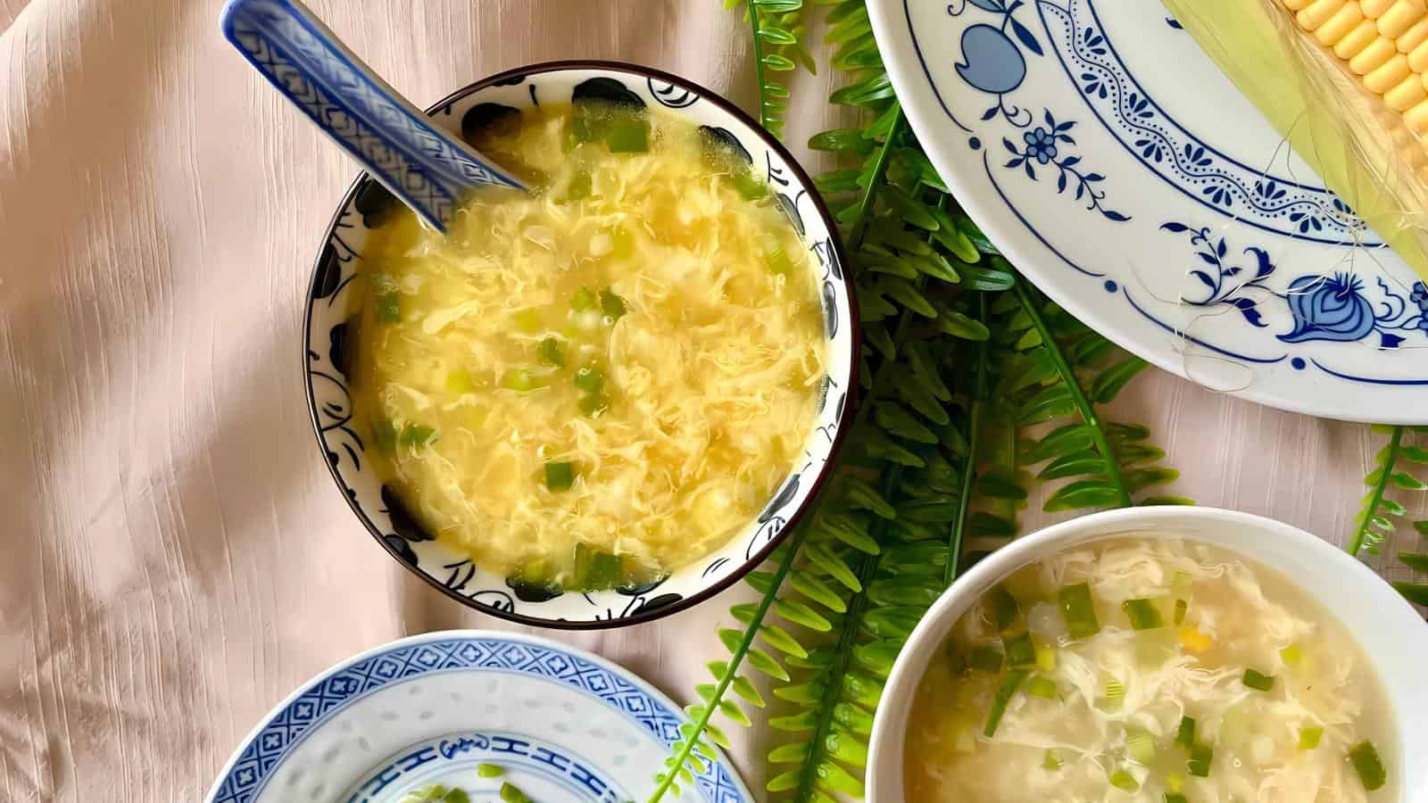 A bowl of egg drop soup with chopped green onions, placed on a table alongside other bowls and a plate of corn, with green fern leaves as decoration.