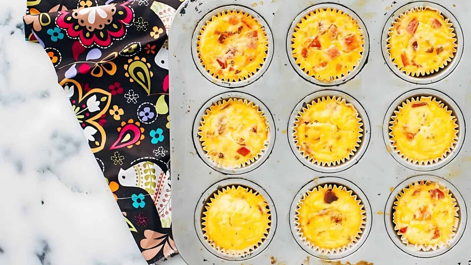 A muffin tin filled with nine baked egg muffins, some with visible bits of vegetables or meat, sits on a marble surface next to a colorful, patterned cloth.