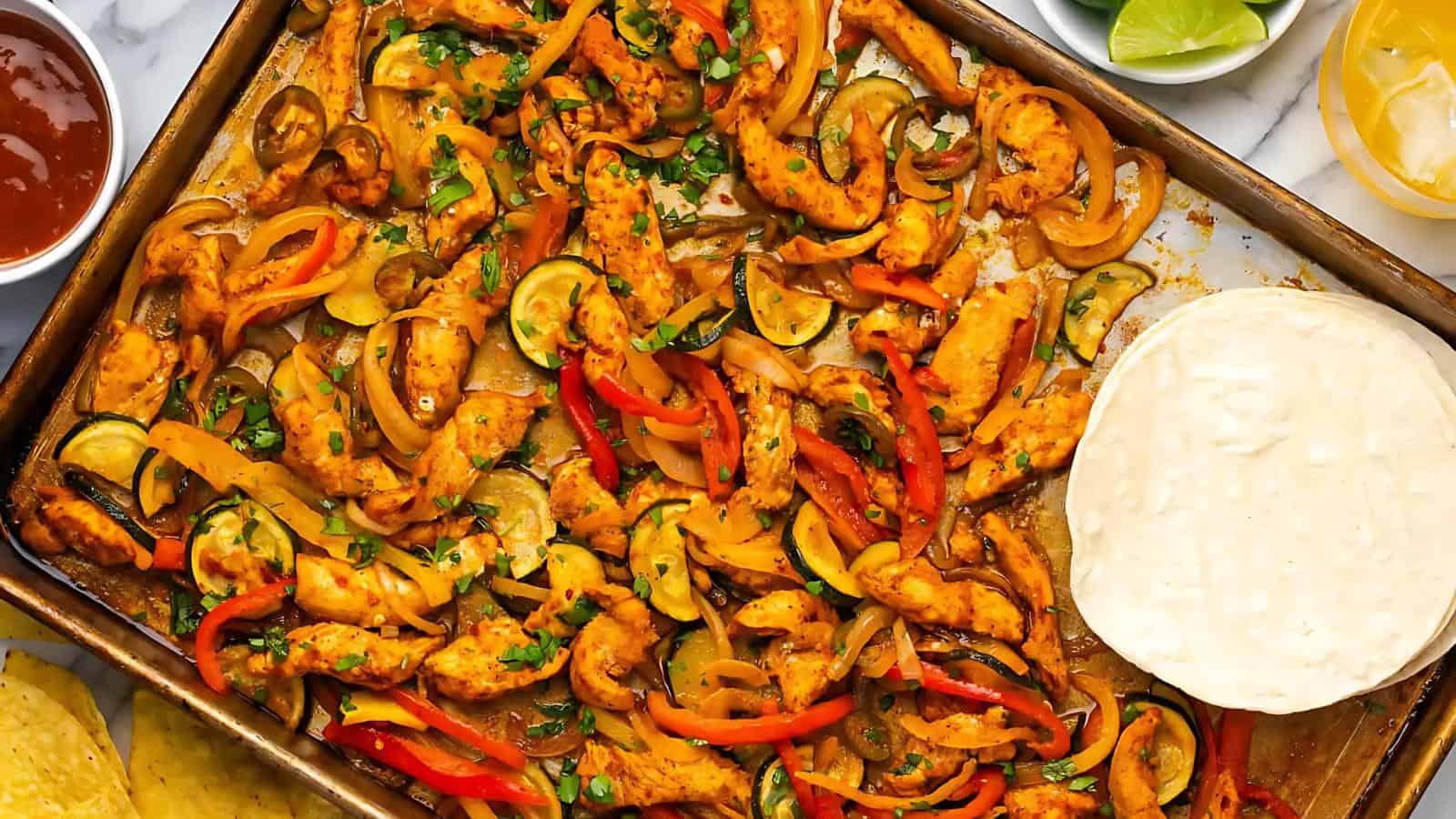 Sheet pan with cooked chicken fajita strips, sliced bell peppers, onions, zucchini, herbs, and a stack of flour tortillas on the side. Lime wedges and salsa are partially visible.