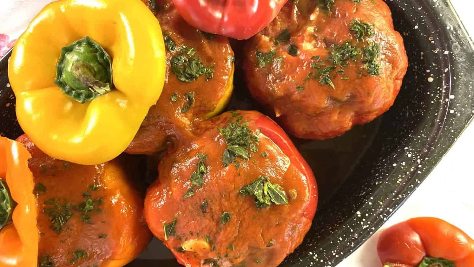 A close-up of stuffed bell peppers topped with tomato sauce and herbs in a black speckled baking dish, with one yellow pepper and several red peppers visible.