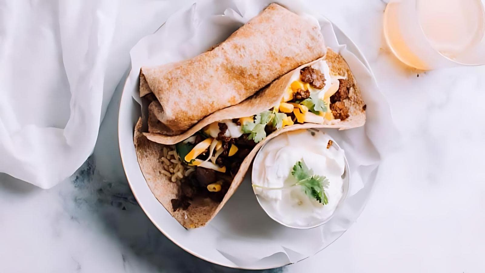 A plate with a burrito and a soft taco filled with meat, corn, and lettuce, served with a side of sour cream and garnished with cilantro. A glass of drink is partially visible.