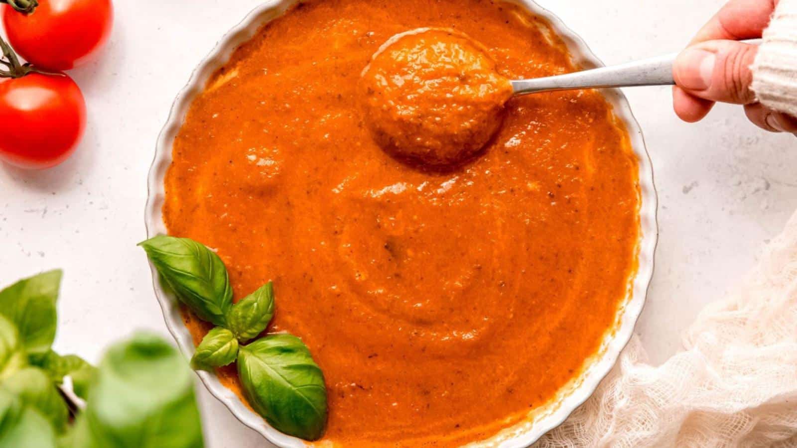A hand holds a spoonful of thick tomato sauce over a bowl, with basil leaves and tomatoes placed nearby on a white surface.