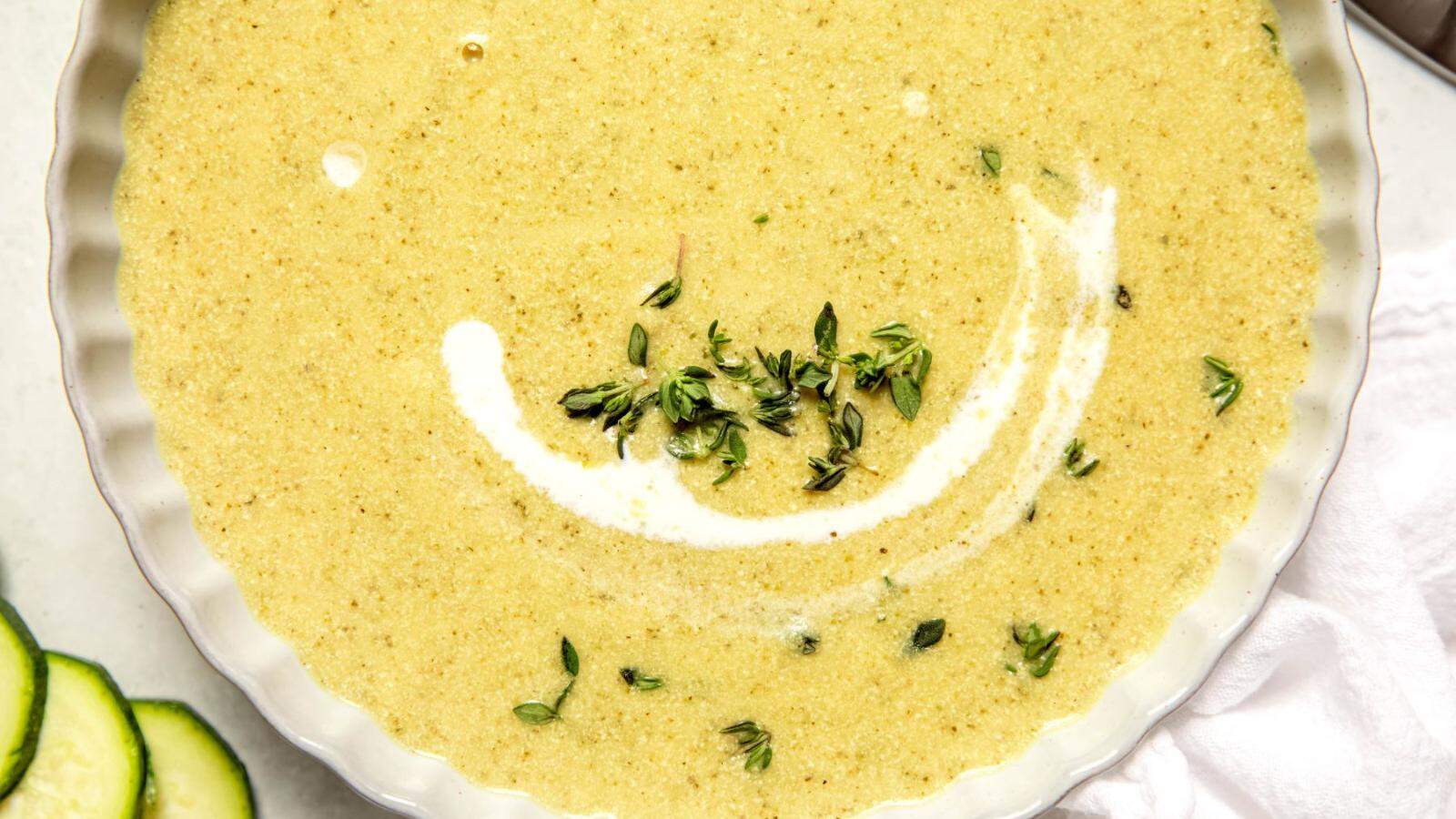 A bowl of creamy green soup garnished with fresh herbs and a swirl of cream, with cucumber slices visible on the side.