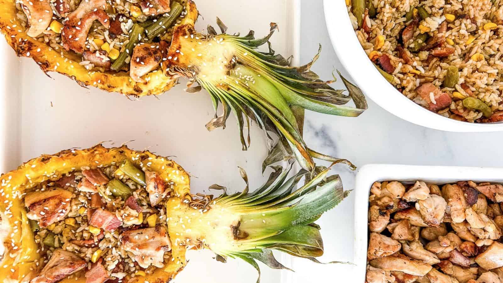 Halved pineapples filled with chicken, vegetables, and rice, alongside bowls of fried rice and cooked chicken pieces on a white surface.