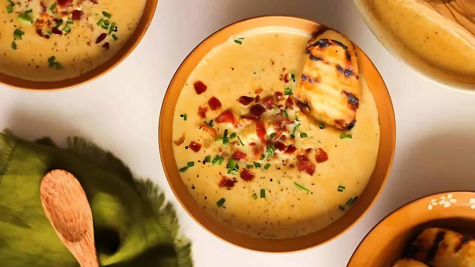 A bowl of creamy soup topped with grilled chicken, chopped chives, and crumbled bacon sits on a white surface next to other bowls and a green cloth napkin.