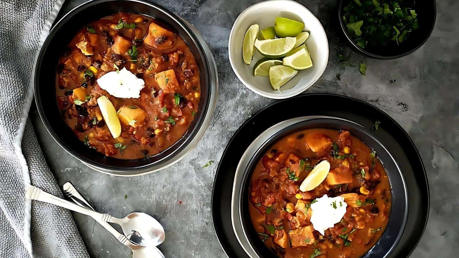 Two bowls of chili topped with sour cream and lime wedges, served with sides of lime wedges, chopped herbs, and two spoons on a gray surface.