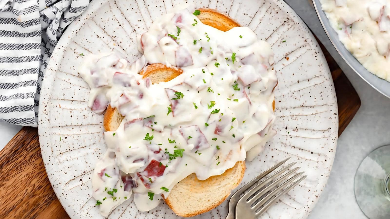 Sliced bread topped with creamy white sauce containing diced meat, garnished with chopped parsley, served on a speckled plate with a fork.