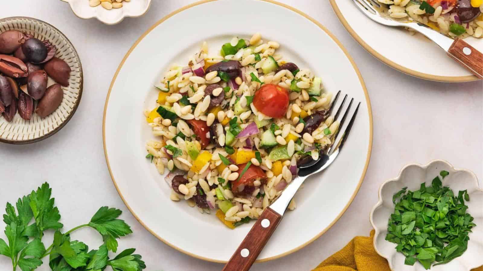 A plate of orzo pasta salad with cherry tomatoes, olives, chopped vegetables, and herbs, surrounded by small bowls of olives and fresh parsley.