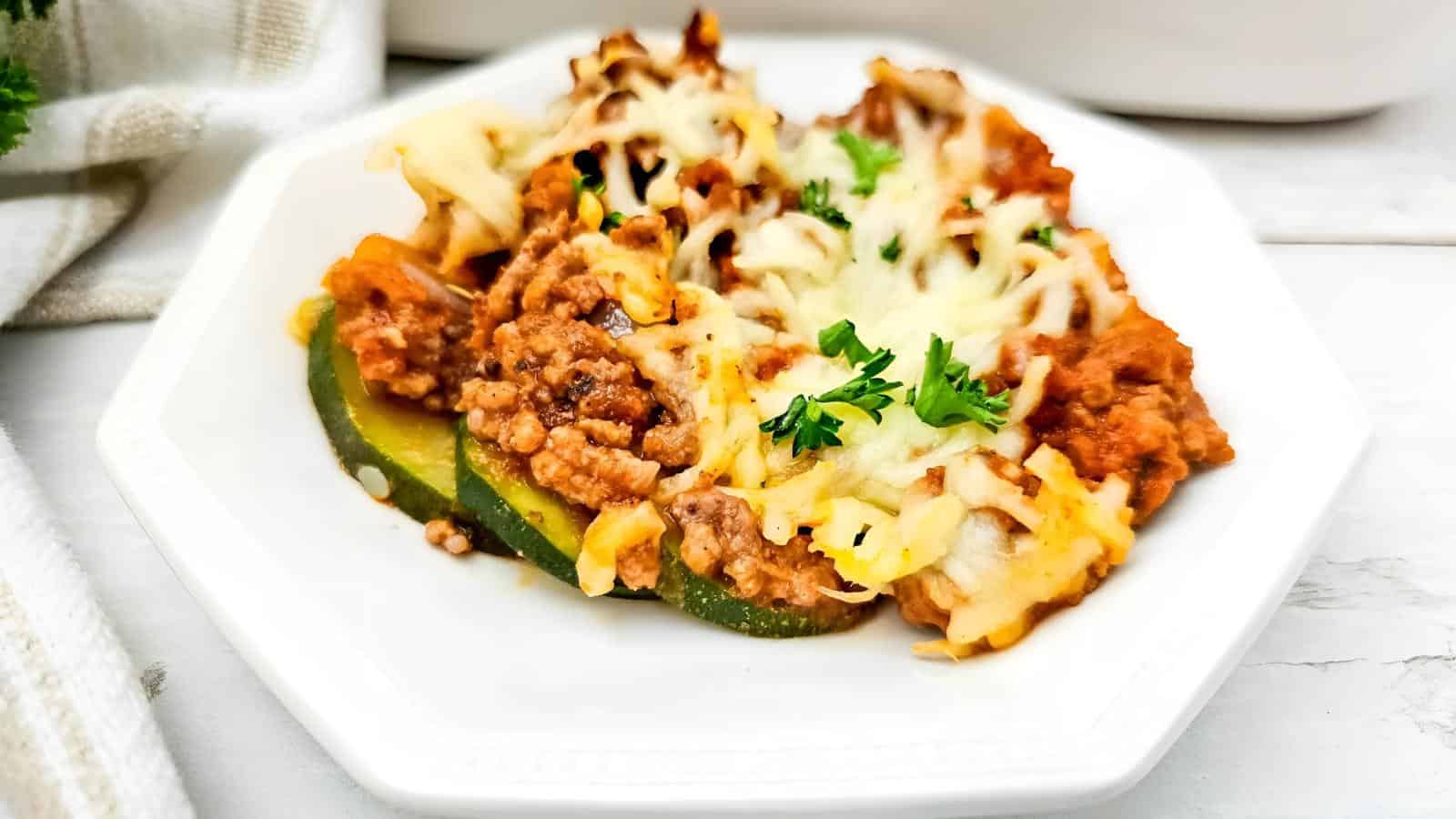 A serving of baked zucchini casserole topped with ground meat, tomato sauce, melted cheese, and parsley on a white plate.