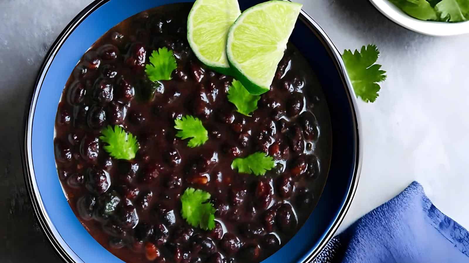 A bowl of black bean soup garnished with cilantro leaves and two lime wedges, placed on a table with a blue napkin and additional cilantro nearby.