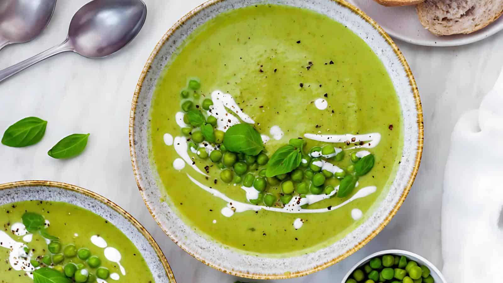 A bowl of green pea soup garnished with whole peas, fresh basil leaves, a swirl of cream, and cracked black pepper, with slices of bread and spoons nearby on a marble surface.