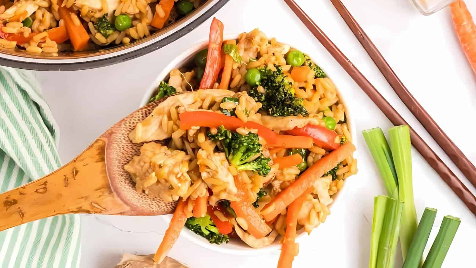 A bowl of vegetable stir-fry rice with carrots, broccoli, peas, and red bell peppers, served with chopsticks on a white surface.