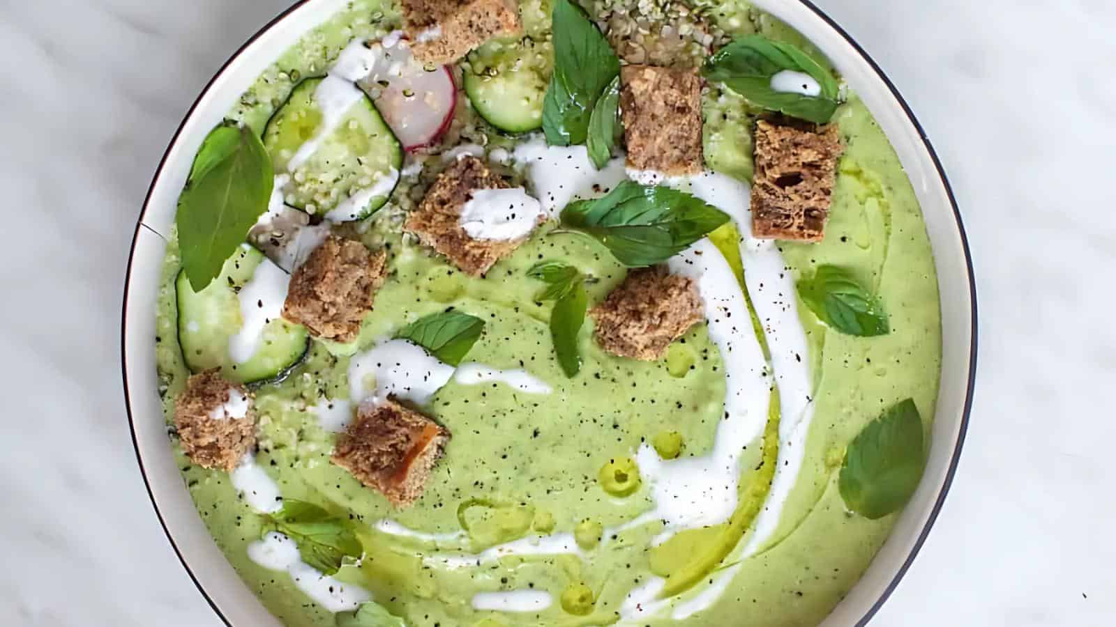 A bowl of green soup topped with croutons, fresh basil leaves, sliced radishes, cucumber, seeds, and a drizzle of cream and olive oil.