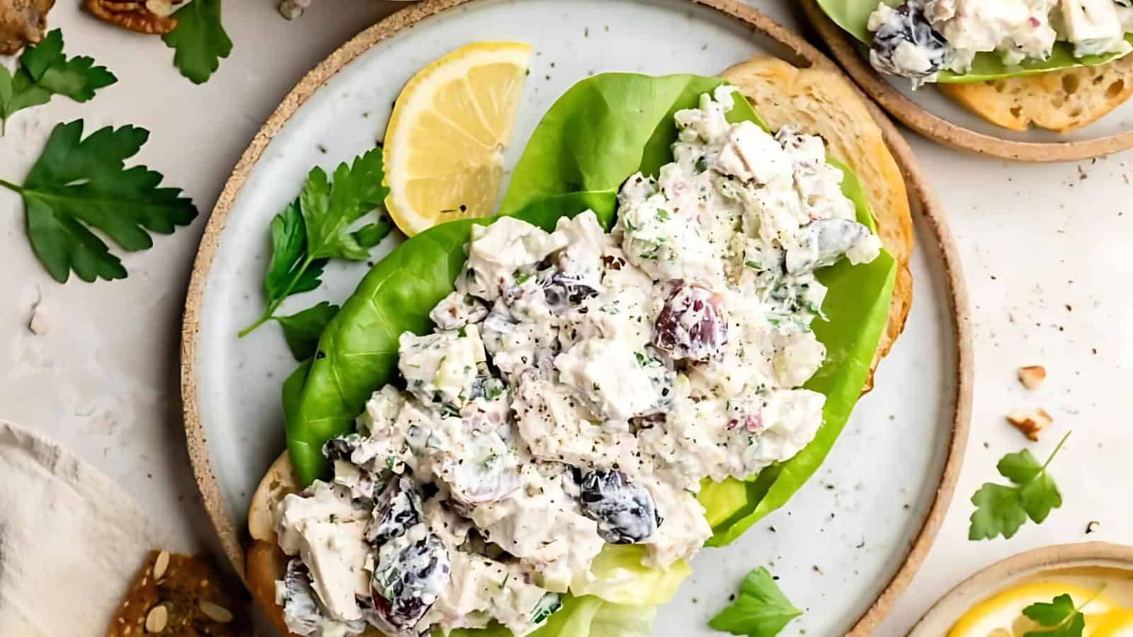 Open-faced chicken salad sandwich with grapes and lettuce on toasted bread, garnished with a lemon slice and fresh parsley on a plate.