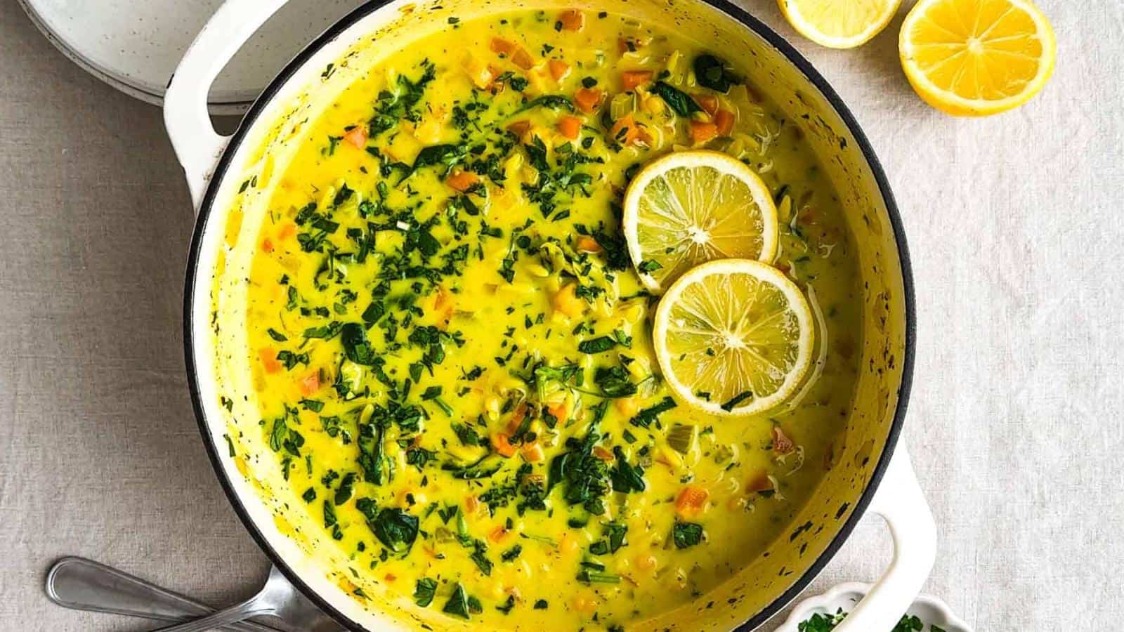 A pot of yellow vegetable soup garnished with chopped herbs and two lemon slices, with halved lemons and plates nearby on a light surface.