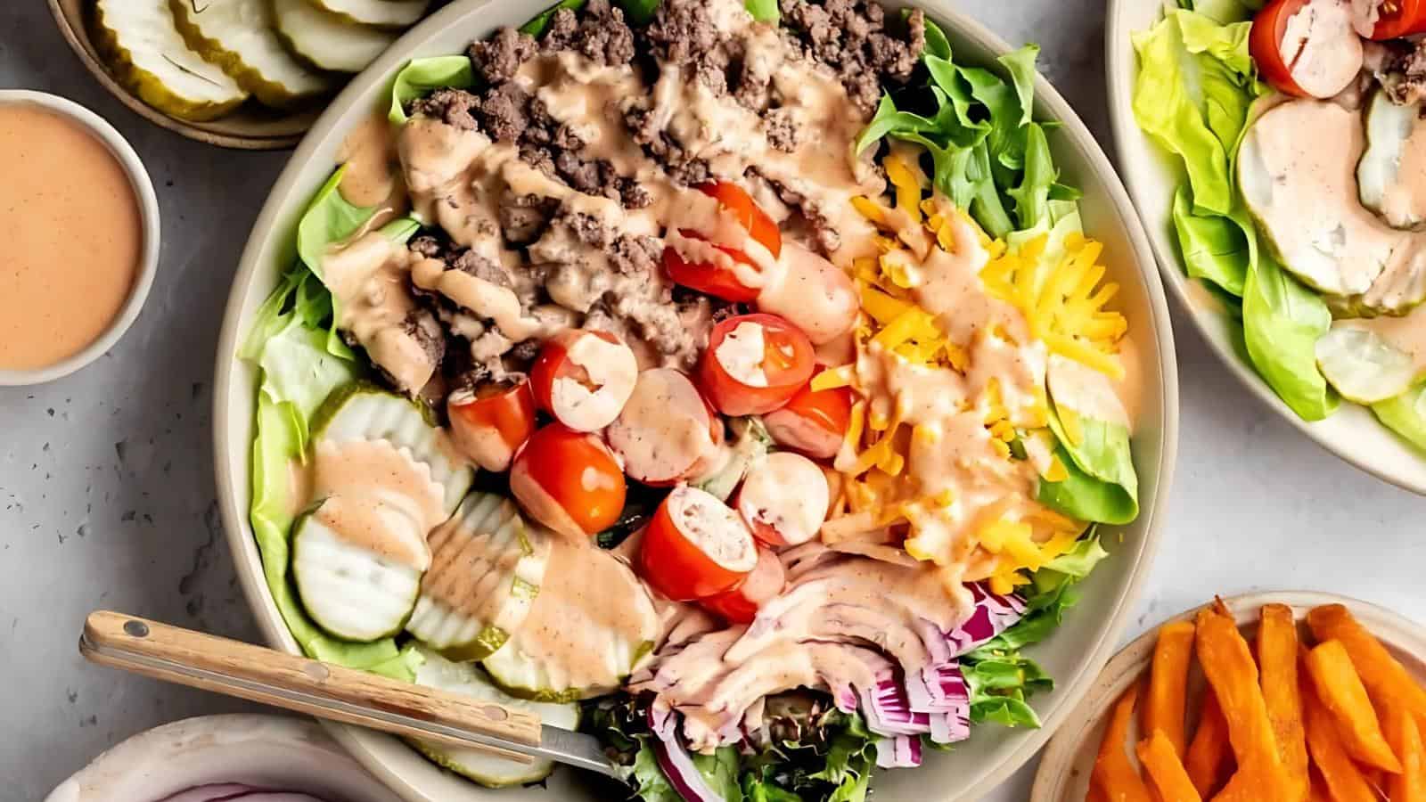 A bowl of salad with ground beef, sliced cherry tomatoes, shredded cheese, pickles, red onions, lettuce, and dressing, served with a fork.