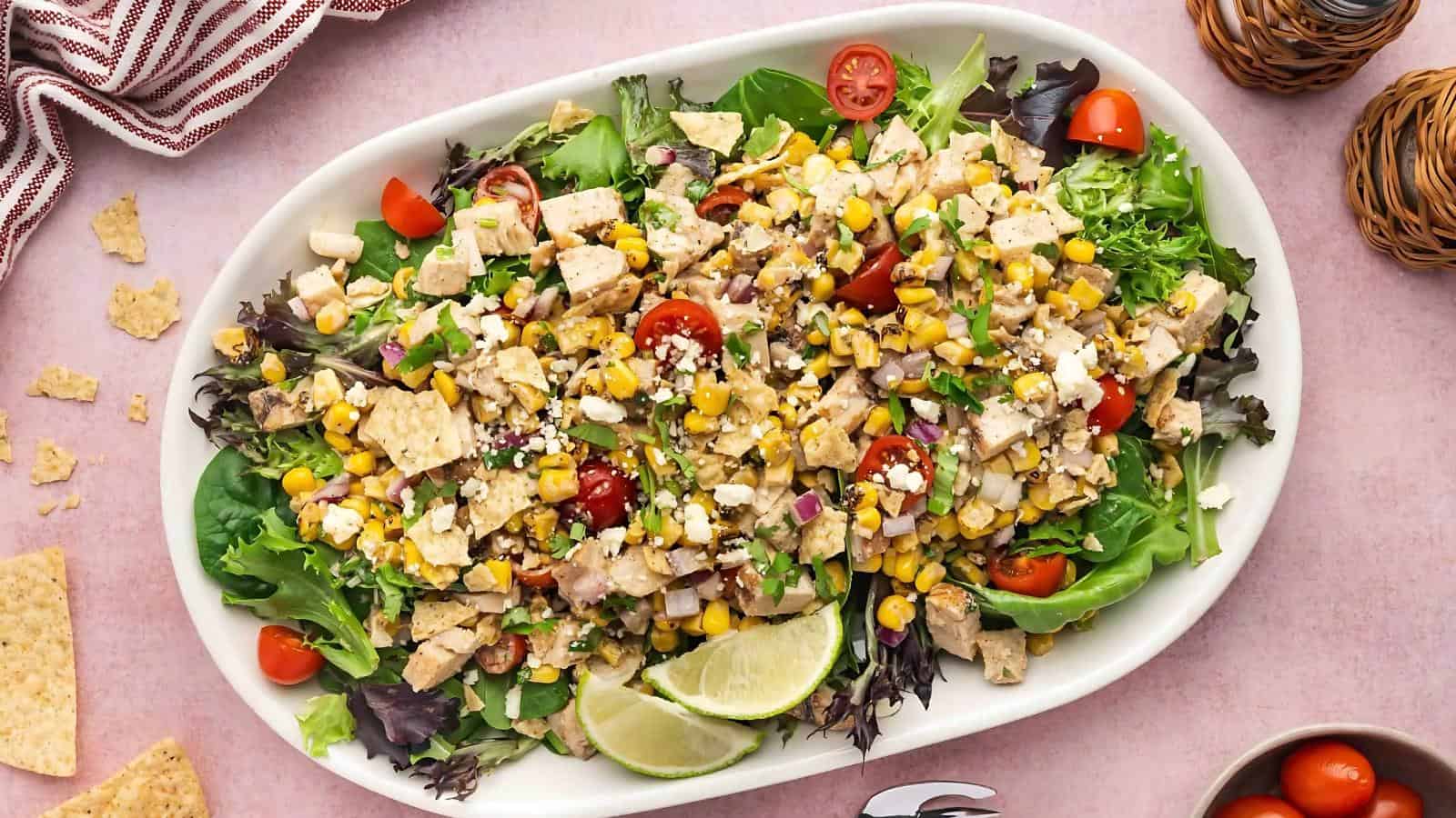 A large white platter filled with chopped salad greens, diced chicken, corn, cherry tomatoes, crumbled cheese, tortilla chips, and lime wedges on a pink surface.