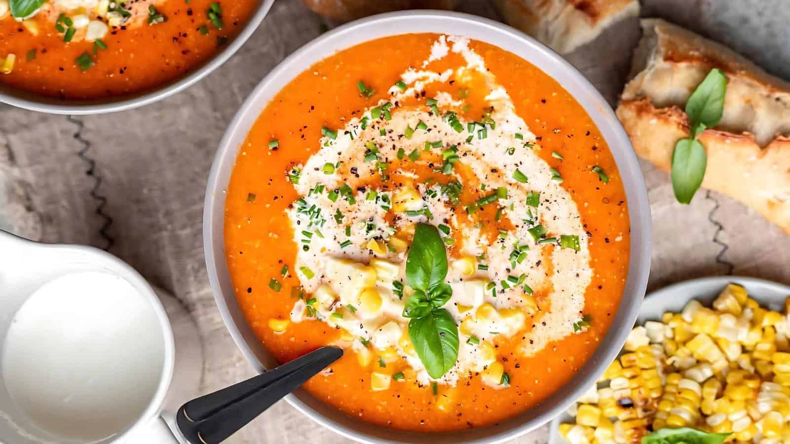A bowl of creamy tomato soup topped with corn, cream, herbs, and a basil leaf, with a spoon in the bowl, bread and corn on the side.