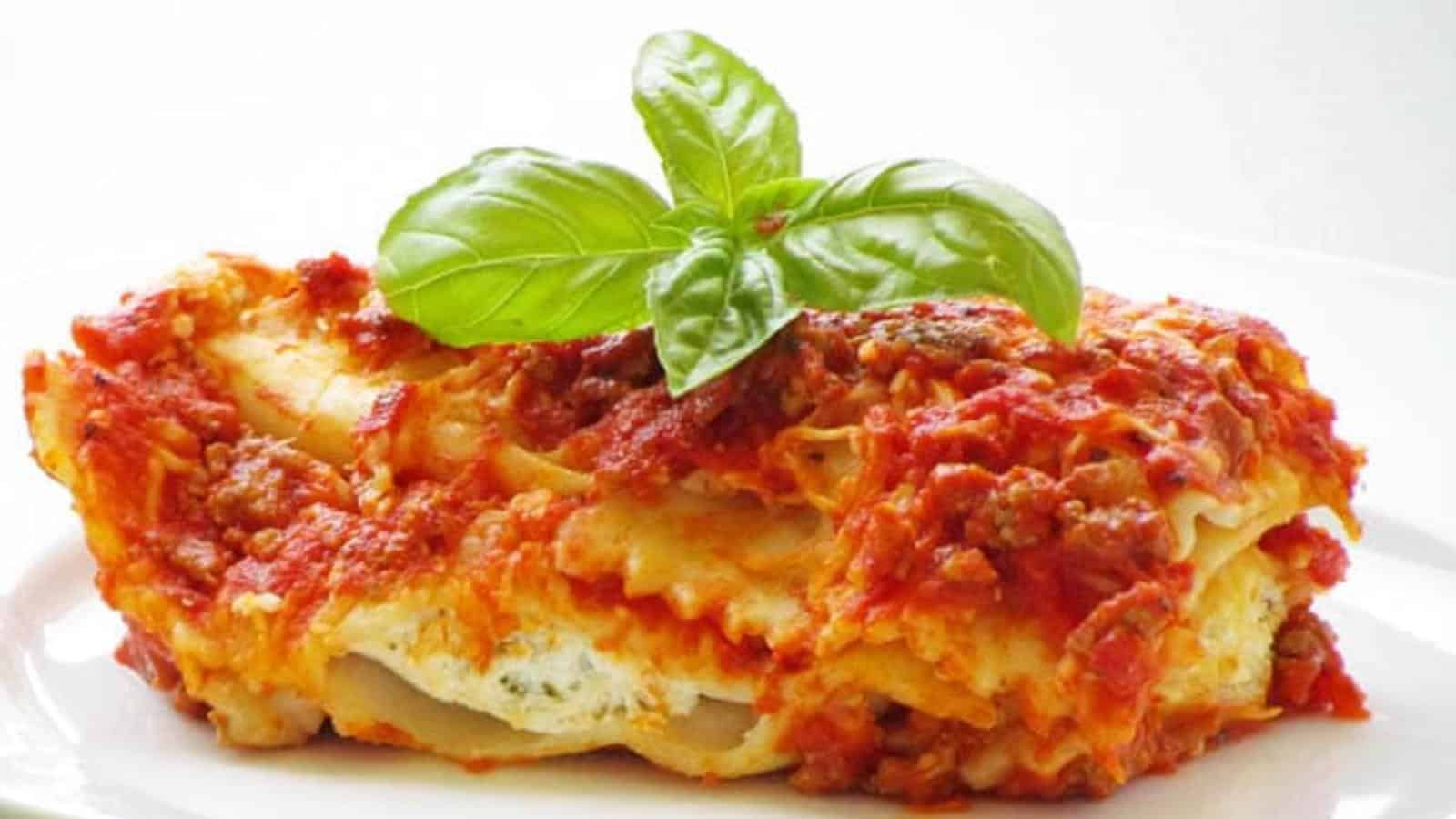 A slice of lasagna topped with tomato sauce, ground meat, melted cheese, and garnished with a sprig of fresh basil on a white plate.