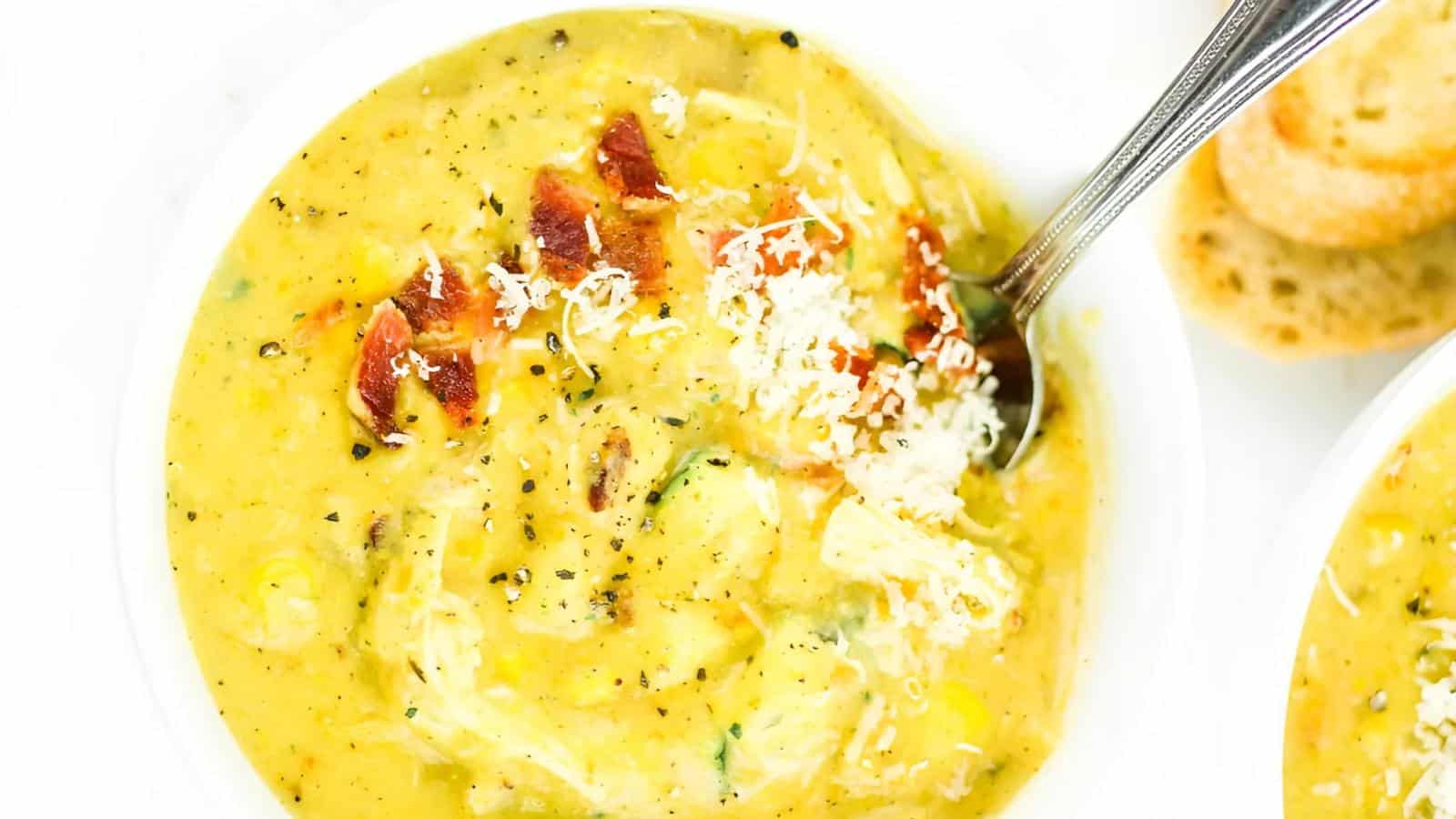 A bowl of creamy soup topped with shredded cheese, bacon pieces, and black pepper, with a spoon in the bowl and slices of bread on the side.