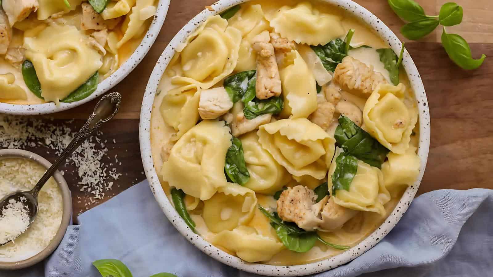 A bowl of cheese-filled tortellini pasta with pieces of chicken and spinach leaves, garnished with grated cheese and served on a wooden table.