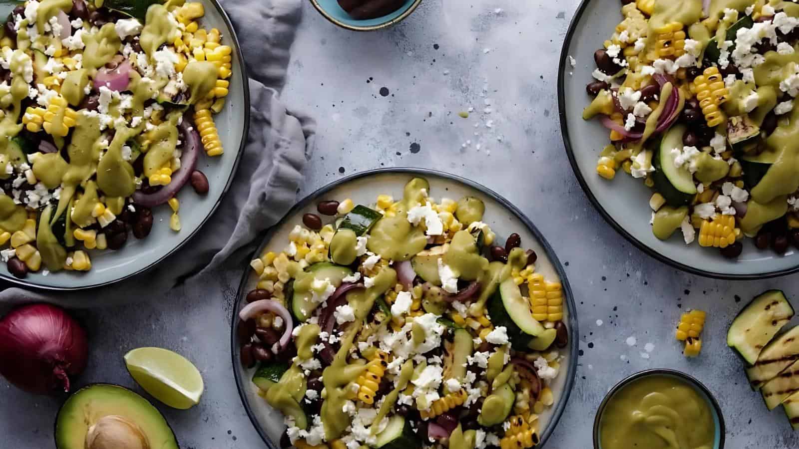 Three plates of salad with corn, black beans, avocado, cucumber, red onion, cheese, and dressing on a gray surface, with lime wedges and extra dressing nearby.