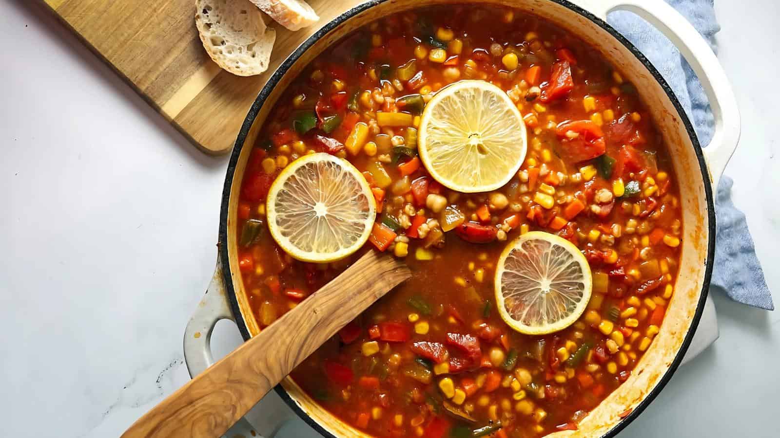A pot of vegetable soup with corn, beans, and tomatoes is garnished with lemon slices. A wooden spoon rests in the pot, and sliced bread is on a cutting board nearby.