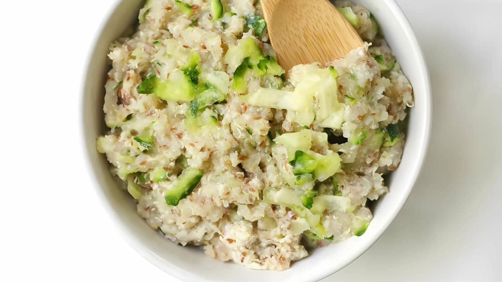 A white bowl filled with a chunky mixture of chopped cucumber and grains, stirred with a wooden spoon.
