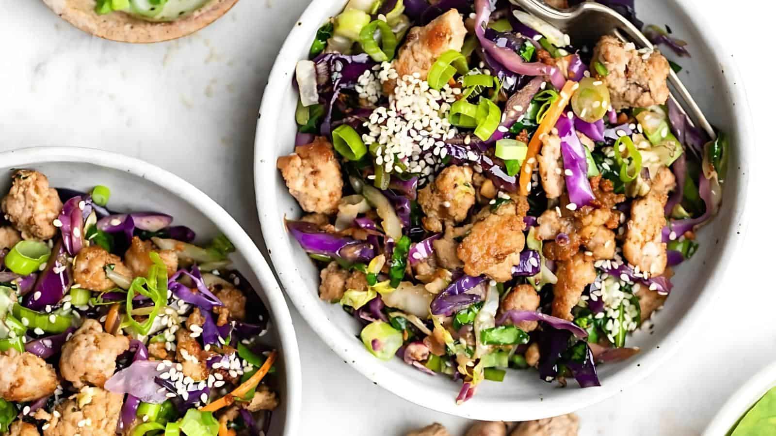A bowl filled with cooked ground meat, shredded purple cabbage, green onions, sesame seeds, and vegetables, with a fork resting on the side.