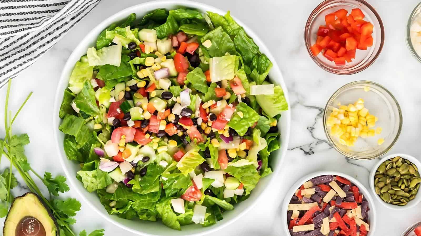 A bowl of chopped salad with lettuce, black beans, corn, tomatoes, onions, and cheese, surrounded by small bowls of ingredients on a white countertop.