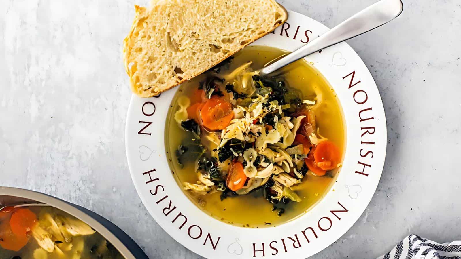 A bowl of chicken and vegetable soup with carrots and leafy greens, served with a slice of bread on a plate that reads "NOURISH.