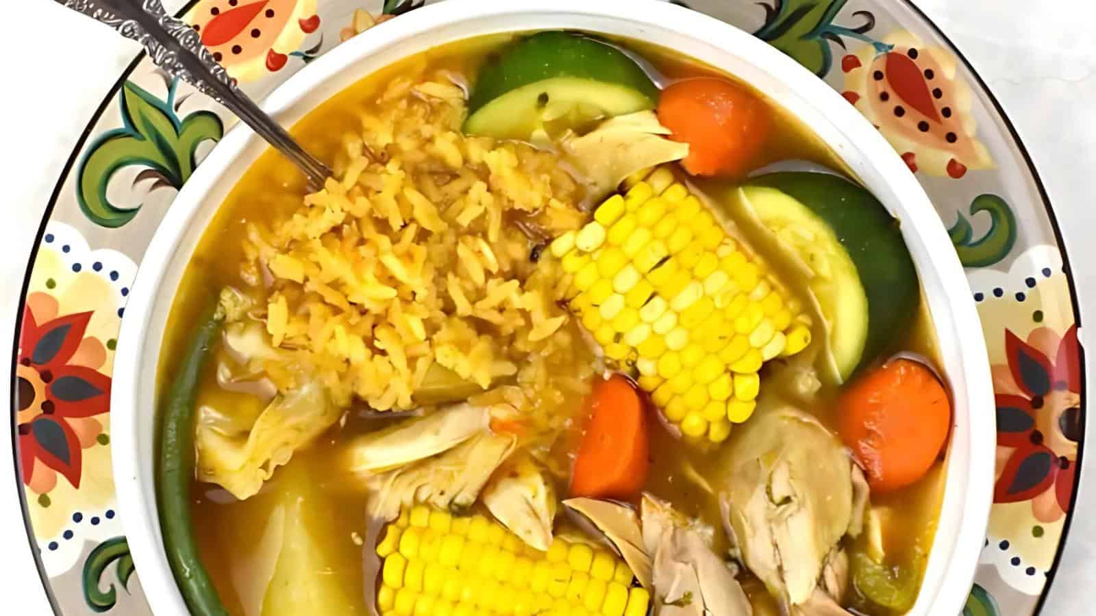 A bowl of soup with chicken, corn on the cob, carrots, zucchini, green beans, and rice, served on a colorful plate.