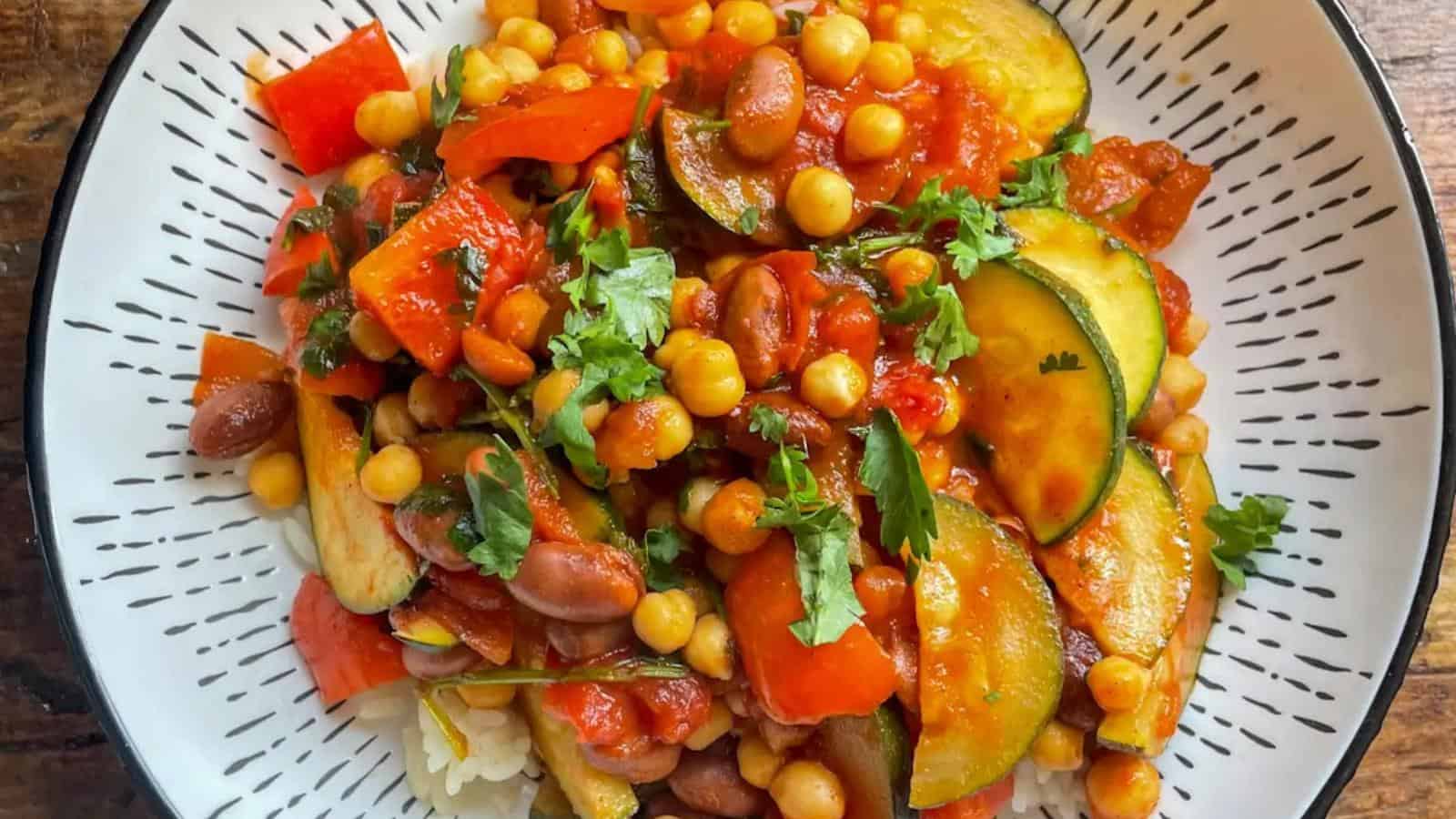 A bowl of rice topped with a colorful vegetable and chickpea stew, including zucchini, red bell pepper, kidney beans, and fresh herbs in a tomato-based sauce.