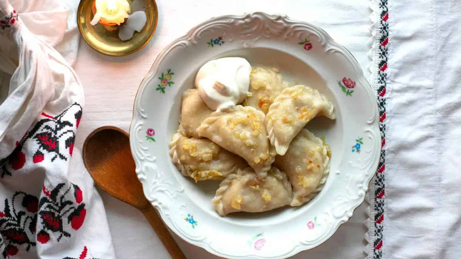 A plate of pierogi topped with sautéed onions and a dollop of sour cream, next to a wooden spoon, candle, and embroidered cloth on a white table.