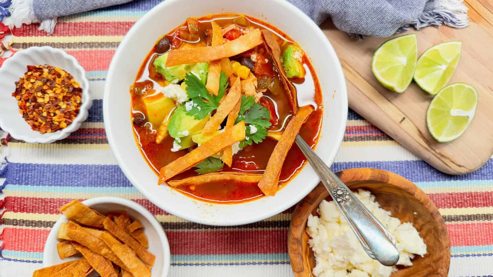 A bowl of tortilla soup topped with avocado, cheese, cilantro, and crispy tortilla strips sits on a striped cloth, with lime wedges, cheese, chili flakes, and extra tortilla strips nearby.