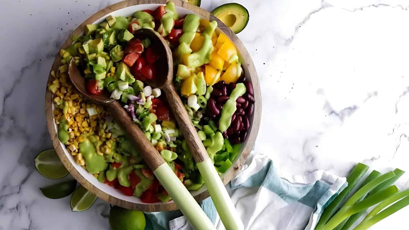A bowl of chopped vegetables, including tomatoes, corn, avocado, beans, onions, and peppers, topped with green dressing, with two wooden spoons on top.