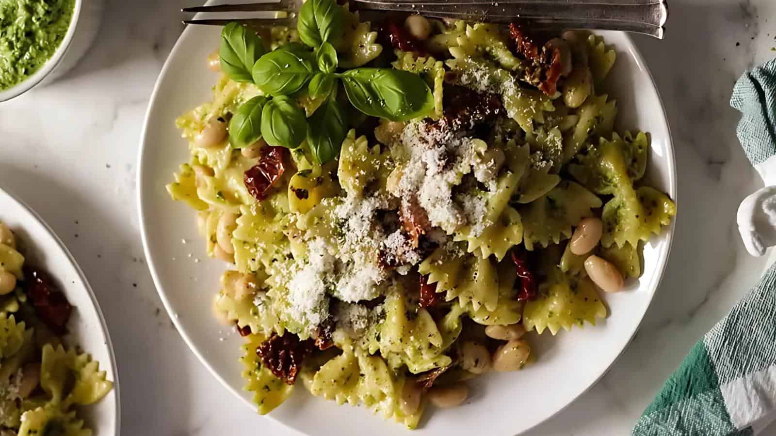 A plate of bow-tie pasta with pesto, white beans, sun-dried tomatoes, grated cheese, and a basil garnish, with utensils on the side.
