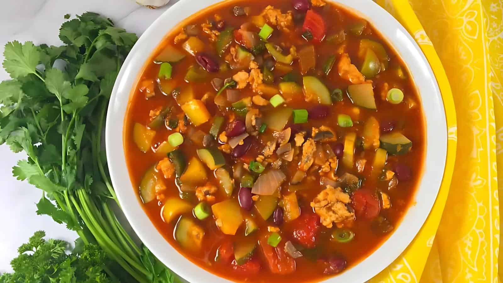 A bowl of vegetable chili with beans, tomatoes, green onions, and diced vegetables, surrounded by fresh herbs on a yellow and white surface.