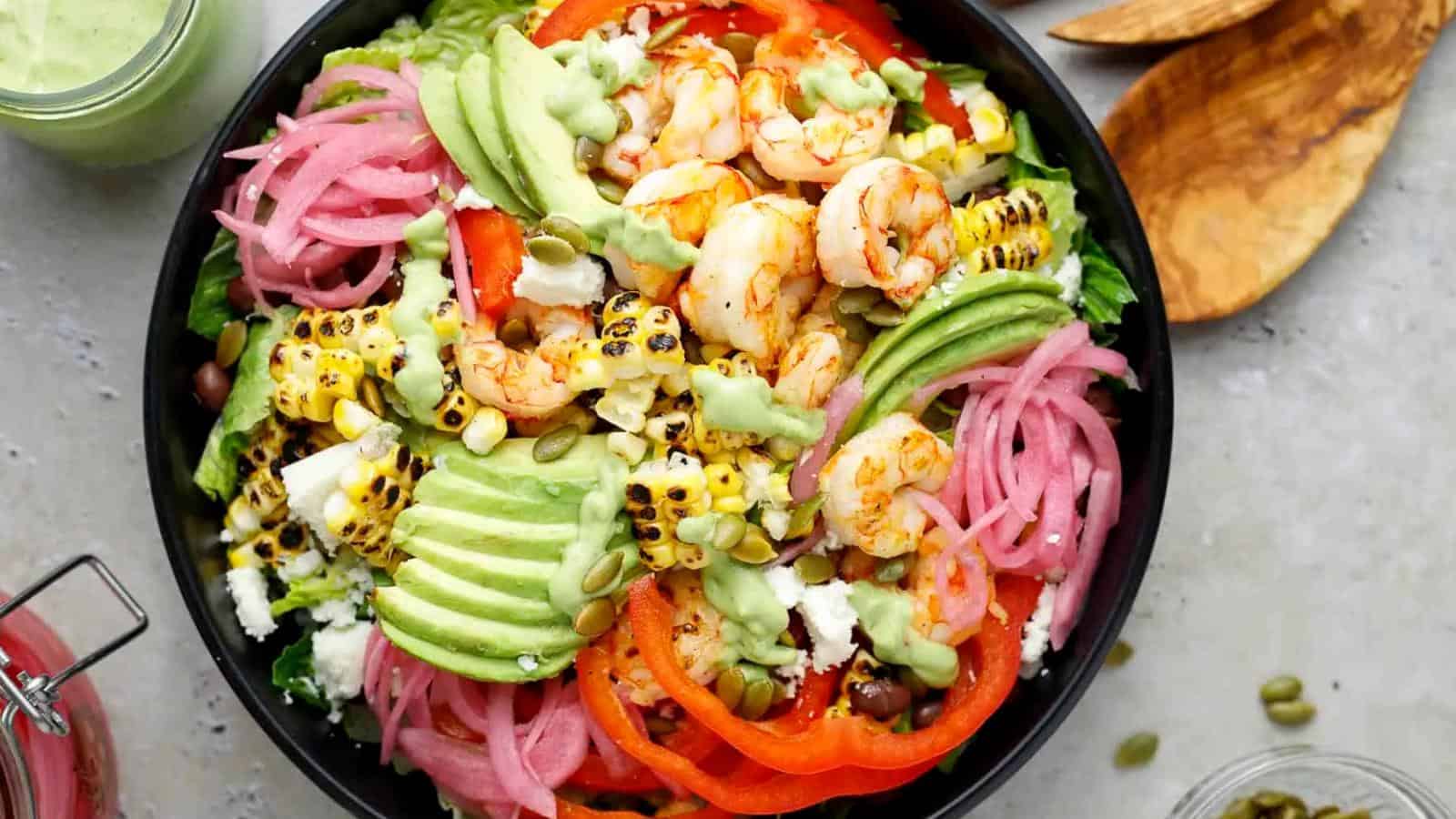 A bowl of salad with grilled shrimp, avocado slices, grilled corn, pickled onions, black beans, tomato, lettuce, pepitas, and green dressing.