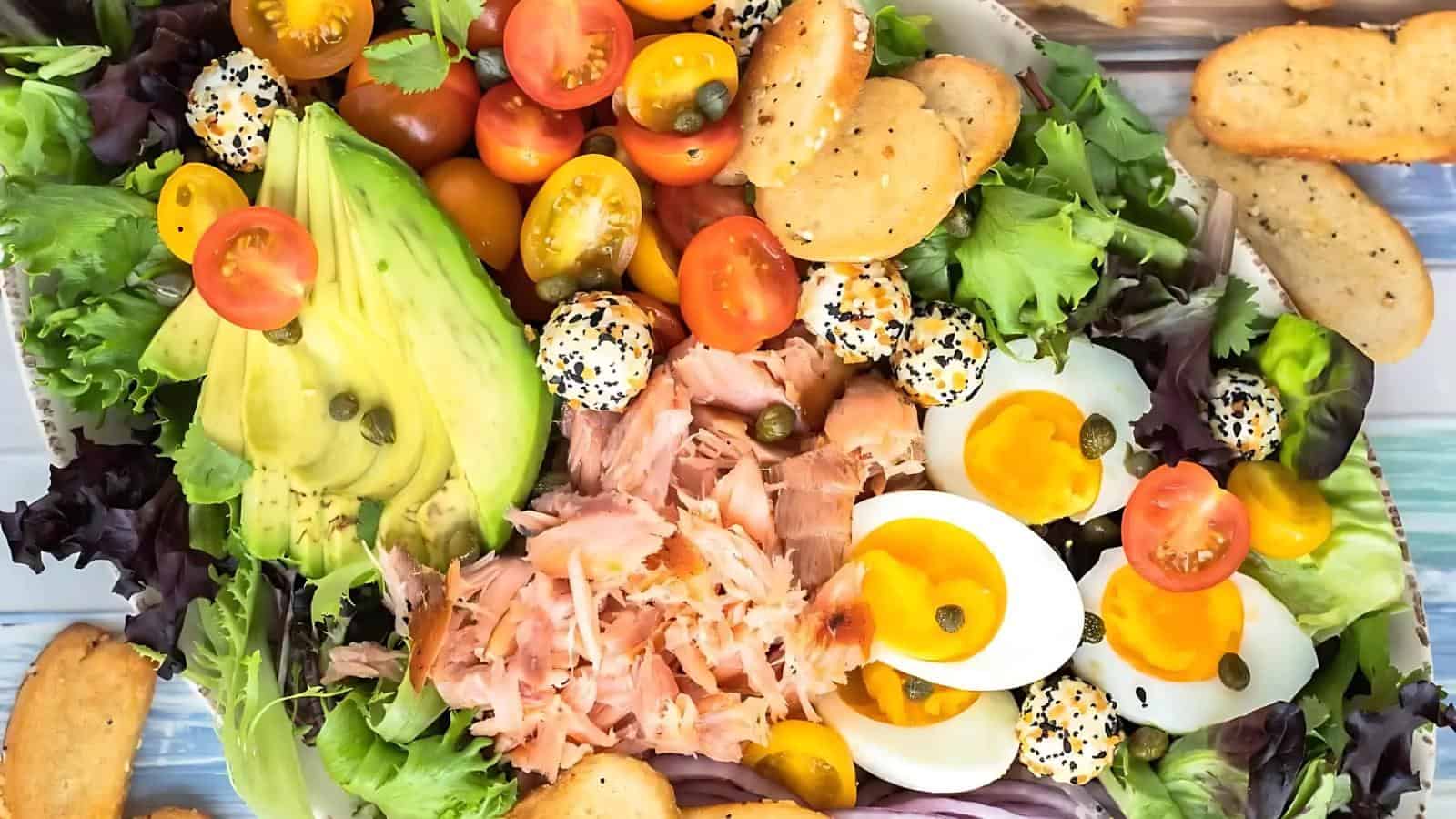 A salad platter with mixed greens, sliced avocado, cherry tomatoes, soft-boiled eggs, smoked salmon, mini cheese balls with seeds, capers, and sliced bread.