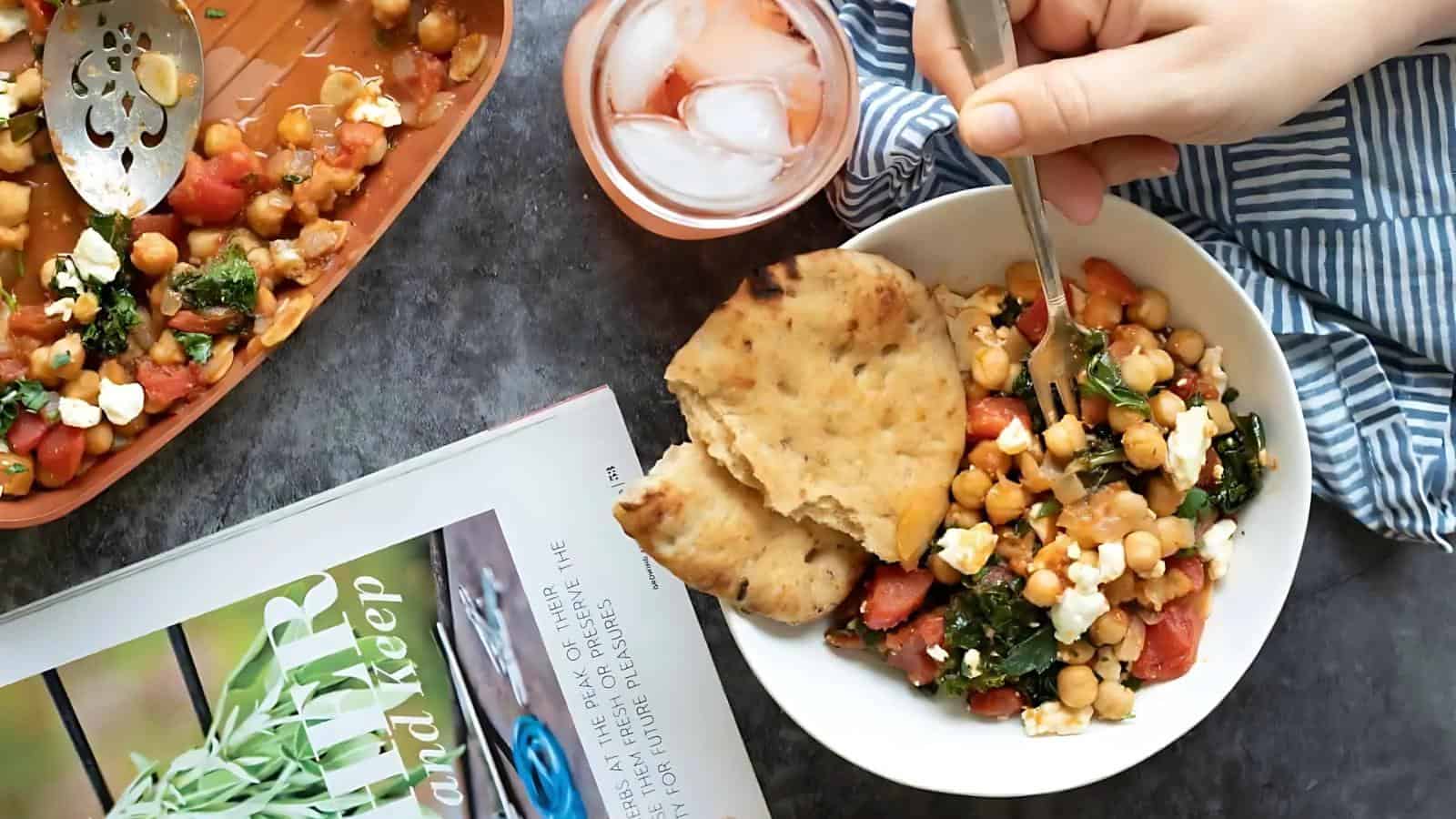A hand holds a fork over a bowl of chickpea salad with naan bread next to a cold drink, a casserole dish, and a cookbook on a gray surface.
