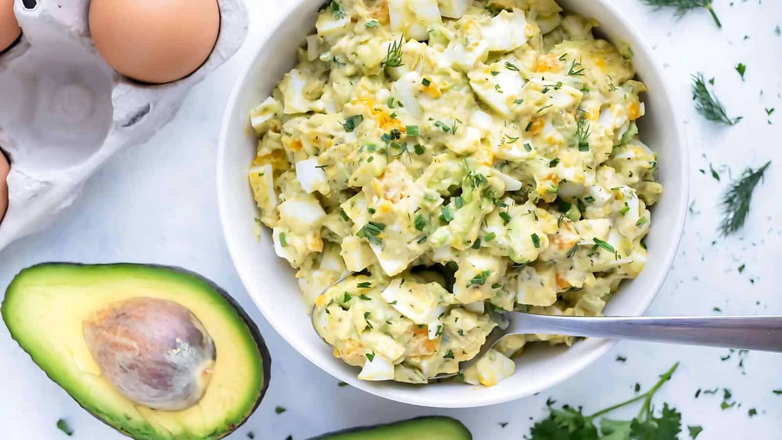 A bowl of egg salad garnished with herbs sits on a white surface near avocado halves, eggs, and fresh herbs, with a spoon in the bowl.
