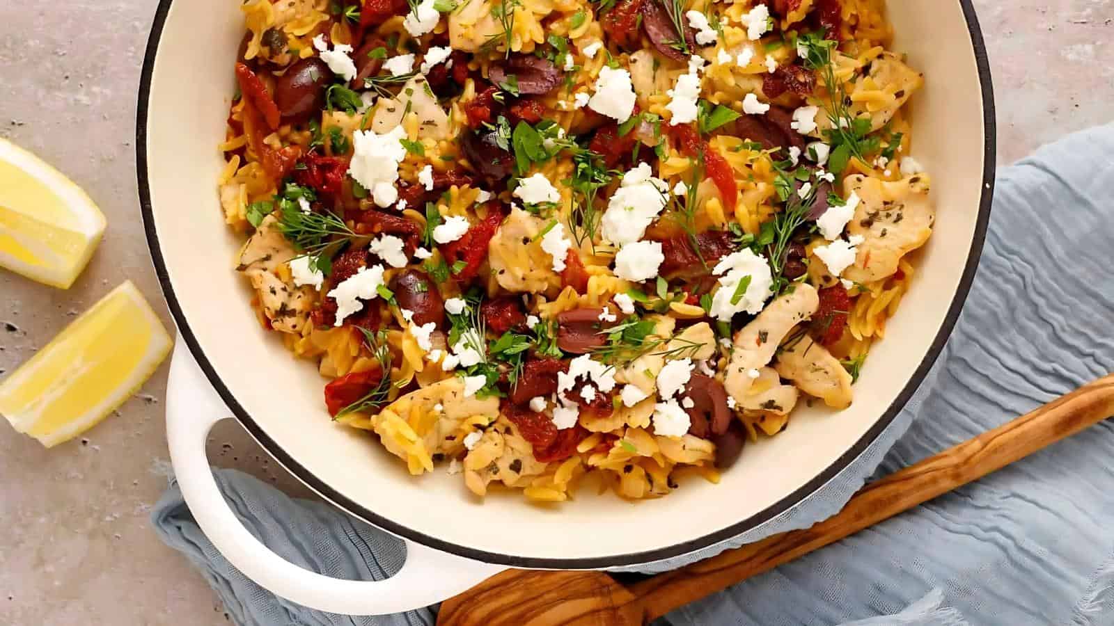 A white pot filled with orzo pasta, chicken, tomatoes, herbs, kalamata olives, and feta cheese, placed on a blue cloth with lemon wedges beside it.