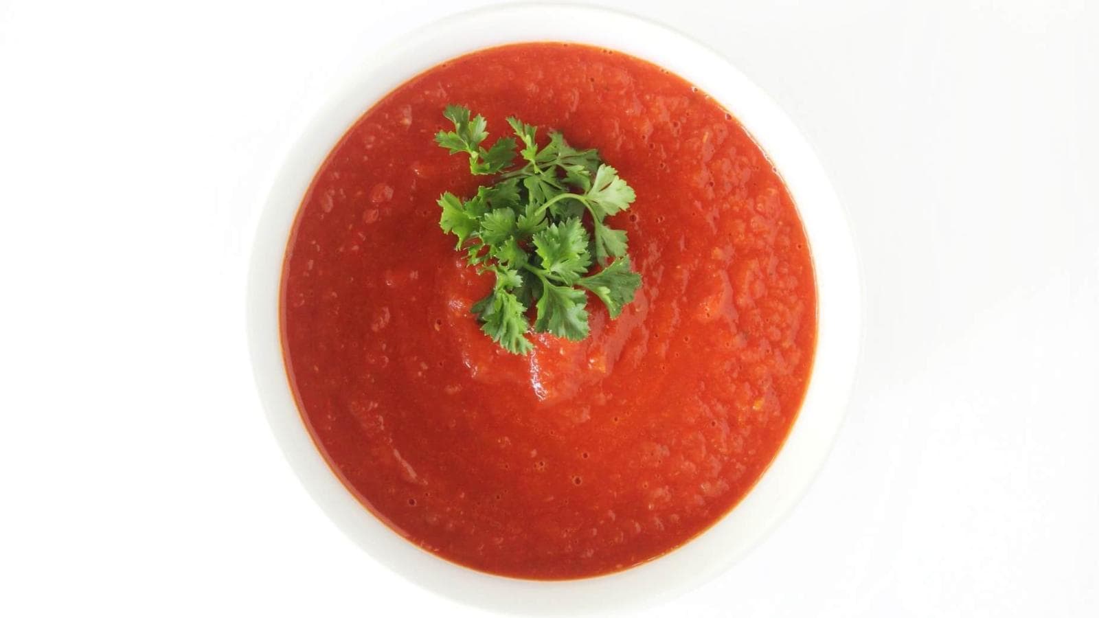 A white bowl filled with red tomato soup, garnished with a sprig of fresh cilantro, viewed from above against a white background.
