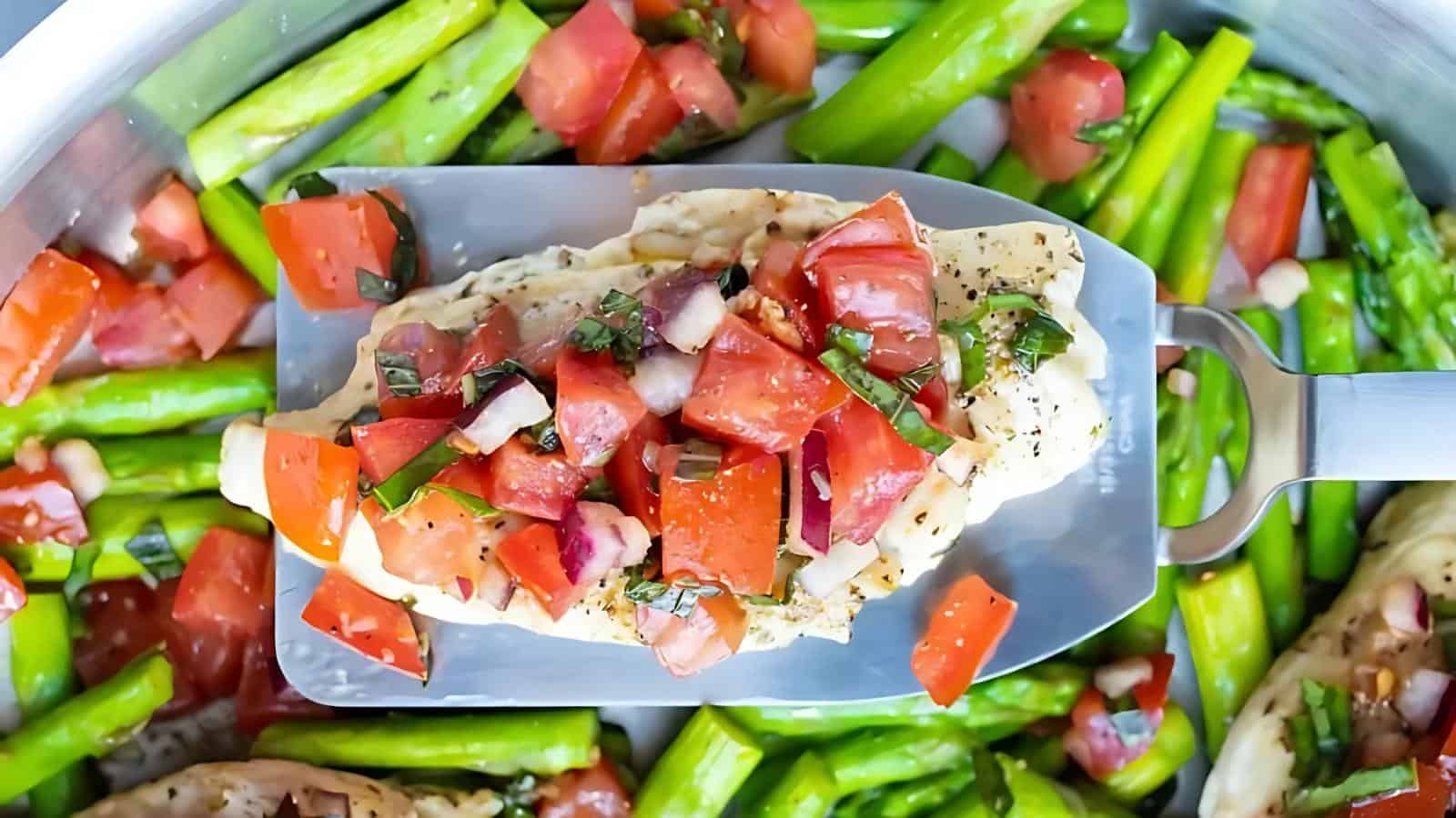 A metal spatula holds a piece of cooked chicken topped with diced tomatoes and herbs over a pan filled with asparagus and more tomato pieces.