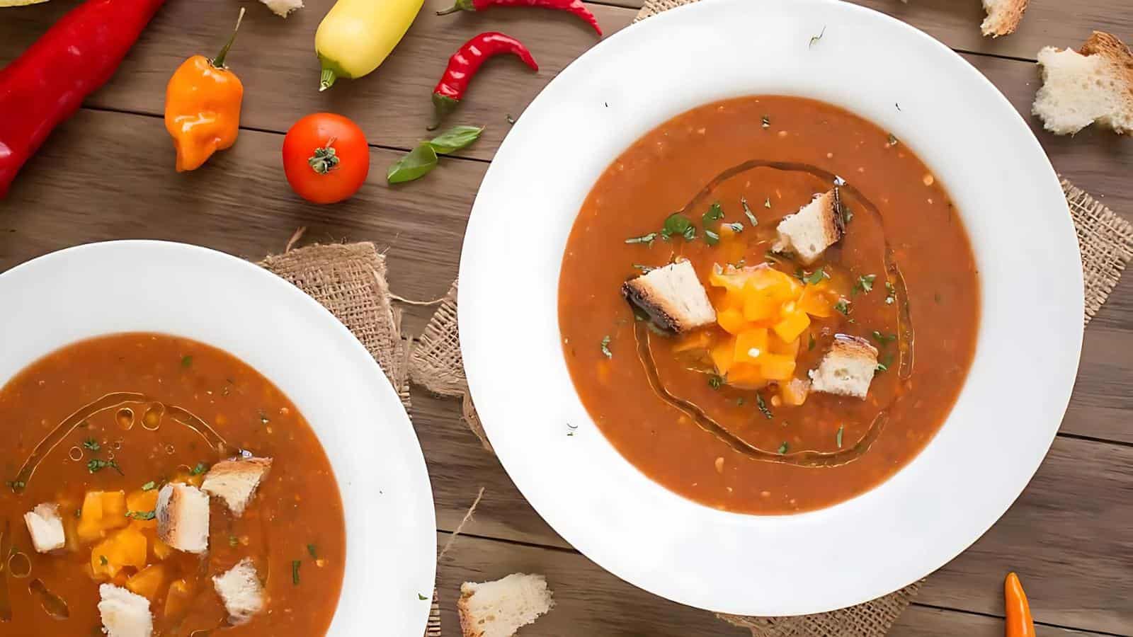 Two white bowls of tomato soup topped with bread cubes and diced yellow bell pepper sit on a wooden table with scattered vegetables and pieces of bread.