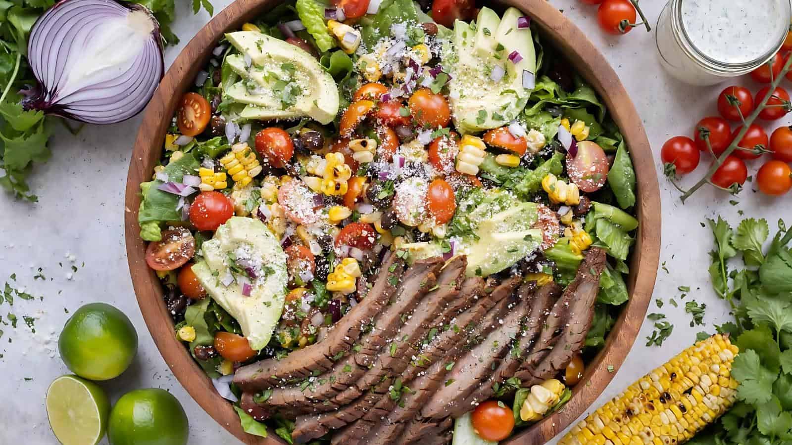 A bowl of salad with sliced steak, avocado, cherry tomatoes, corn, red onion, lettuce, and cheese, garnished with herbs. Fresh limes, tomatoes, and corn surround the bowl.