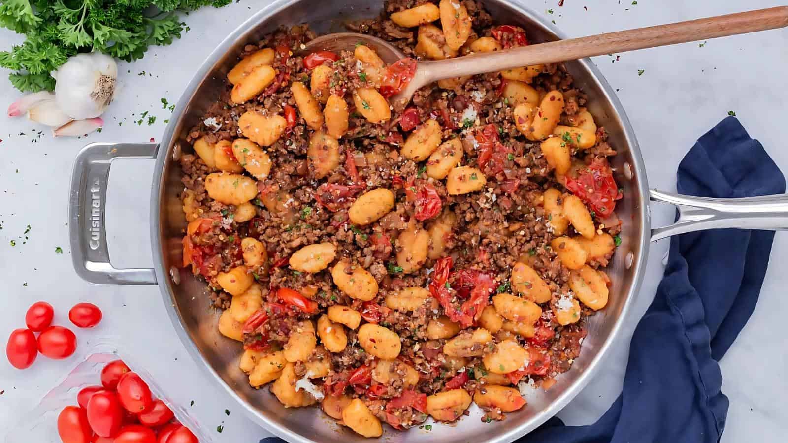 A stainless steel pan filled with gnocchi, ground meat, tomatoes, and herbs, with a wooden spoon resting in the food.