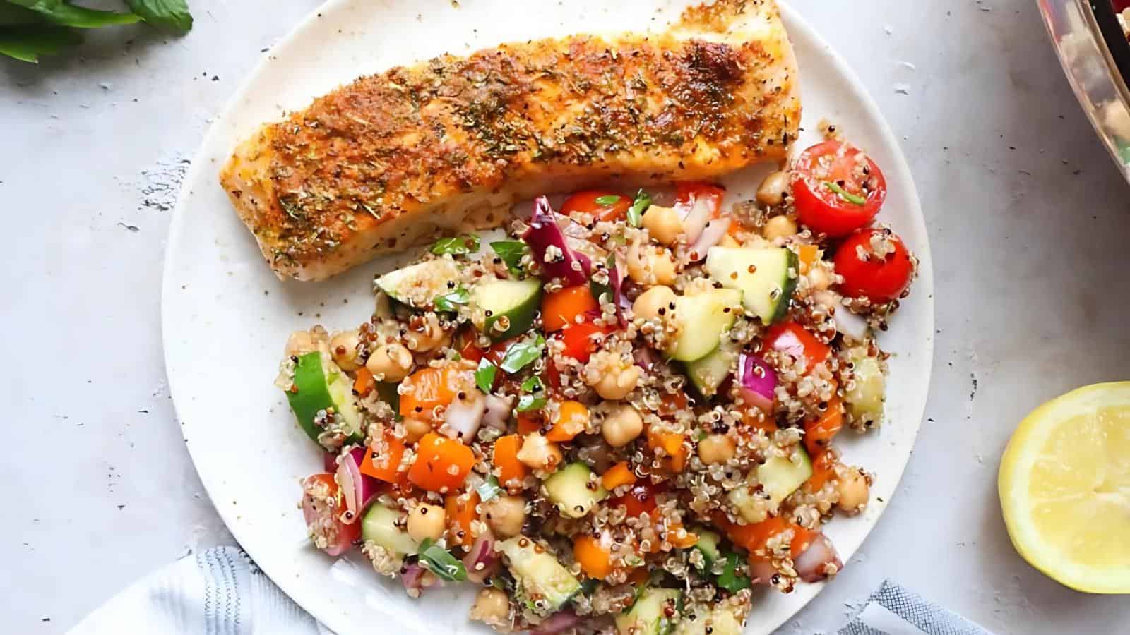 A plate with a seasoned, baked salmon fillet and a colorful quinoa salad with chickpeas, cherry tomatoes, cucumber, red onion, parsley, and orange bell pepper.